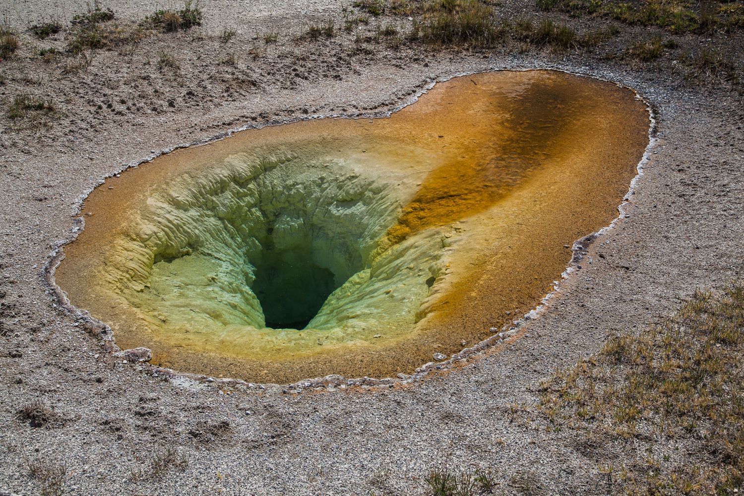 An oval hot spring set in flat gray gravel. The near half is a funnel-shaped opening with ribbed yellow-green walls dropping into dark green water at the center. The far half is a shallow shelf covered in orange and mustard-colored mat under clear water. A thin crust of white mineral rims the whole pool. Sparse dry grass grows at the edges.