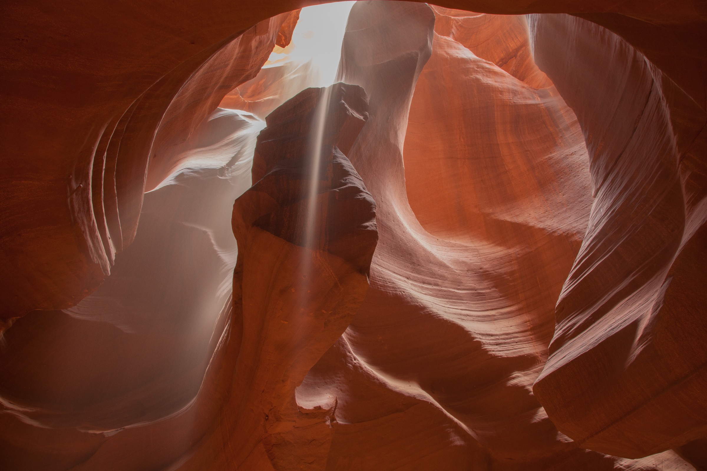 The inside of a narrow, deep canyon in orange and red sandstone, photographed looking upward. The walls curve and fold around the frame in smooth, rounded, layered surfaces, banded light and dark along the bedding, with no sharp edges. At the top center a narrow gap opens to a bright white sky. A single beam of light angles down from that gap through the middle of the frame, and fine particles are visible falling and drifting within it. The lower left of the frame is hazy with suspended dust; the right-hand walls are lit warm orange.