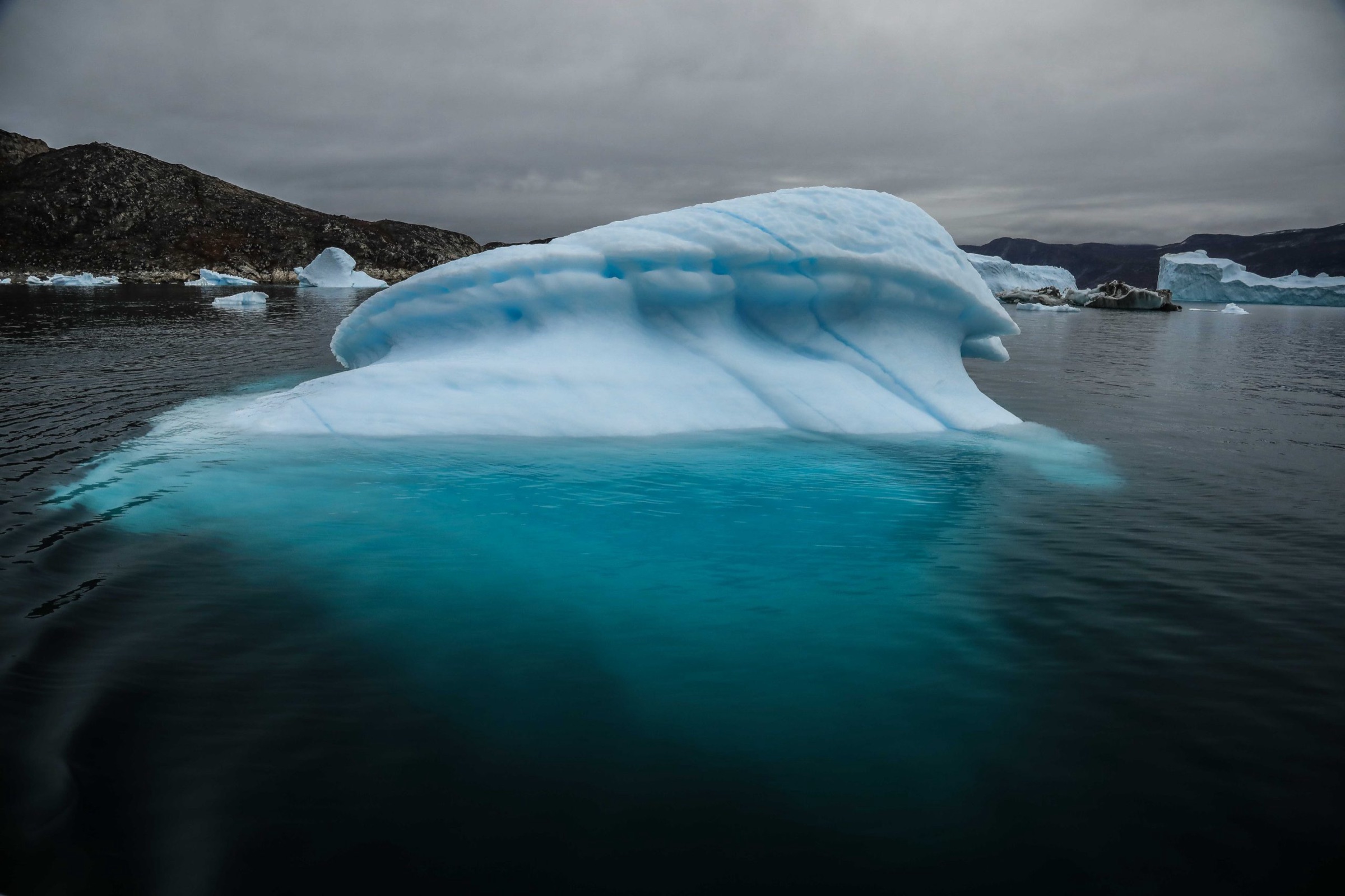A large blue-white iceberg looms over dark water near Ilulissat Icefjord, Greenland, with rocky hills and an overcast sky behind it.