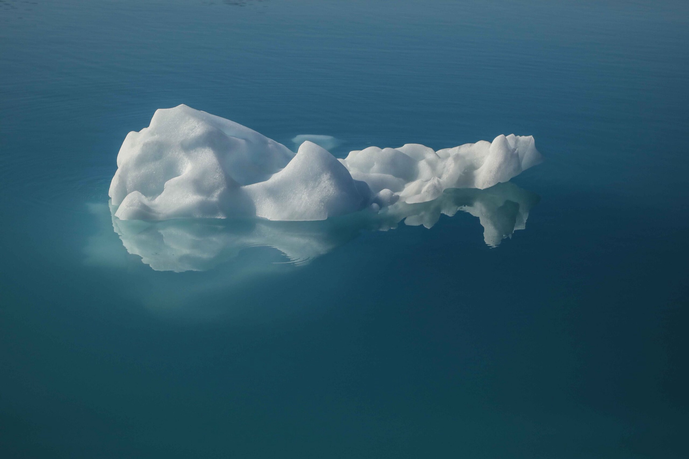 A small iceberg fragment floats on calm, deep blue water off Greenland, its submerged form visible as a pale reflection beneath the surface.